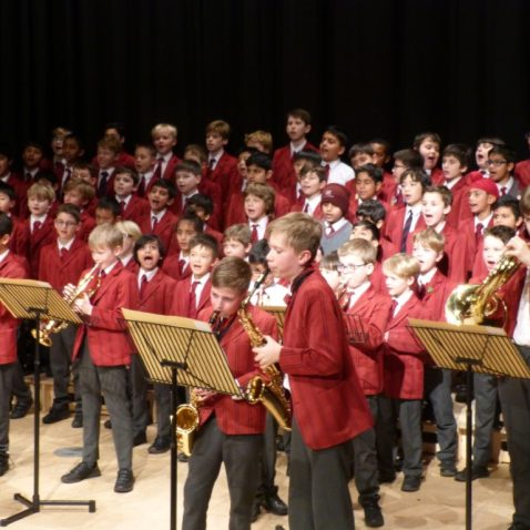 Child playing on the saxophone as a big group of students sing and play instruments