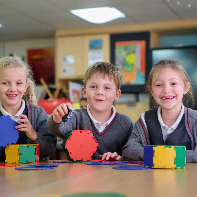 Two girls and a boy in their grey school uniform are sitting in a classroom. They are all smiling and have made constructions from interlocking colourful plastics