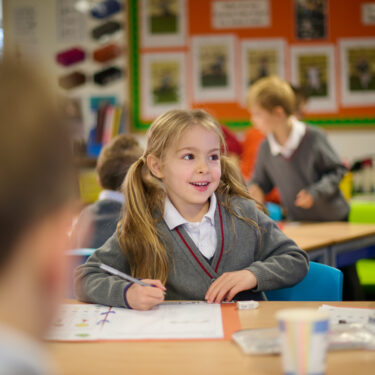 Girl with bunches is looking up and smiling. She's holding a pen and about to write in her schoolbook
