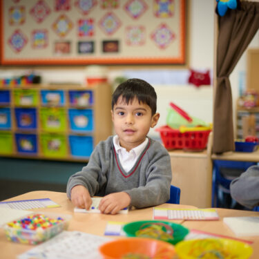 Dark-haired schoolboy in grey jumper is sitting at a table in a classroom and making a pattern with coloured beads