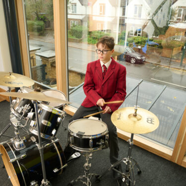 Schoolboy sitting on a stool in front of a drumkit, playing the cymbal and drums. He's in front of a window which looks out onto the wet road