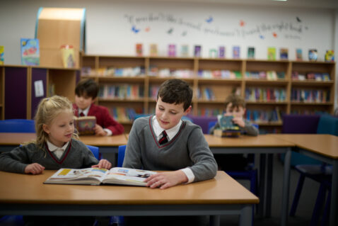 Older boy pupil with younger girl, both in the library reading a colourful book