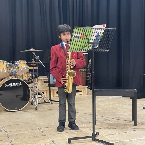 Schoolboy playing saxophone in front of a music stand. There's a drumkit behind him