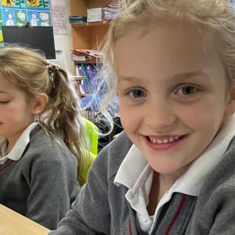 Blonde-haired schoolgirl smiling at the camera. She's sitting in a colourful classroom