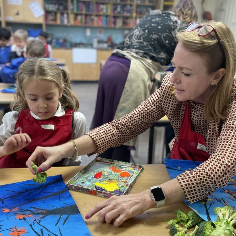 Teacher sits with schoolgirl and shows her how to use broccoli as a paint brush to add leaves to a picture of trees silhouetted against the night sky